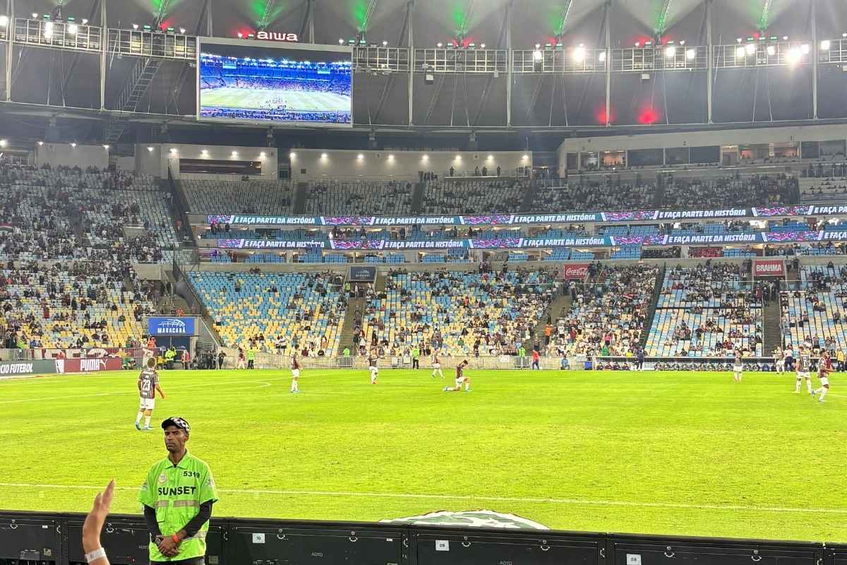 Jogadores do Fluminense em campo no Maracanã vistos da beira do gramado, com arquibancadas e telão ao fundo