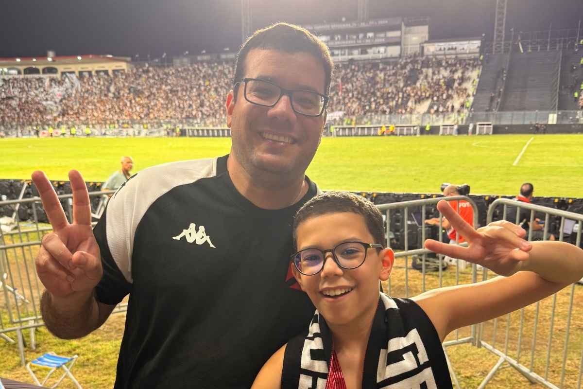 Pai e filho torcedores do Vasco sorrindo e fazendo sinal de vitória com o gramado de São Januário ao fundo